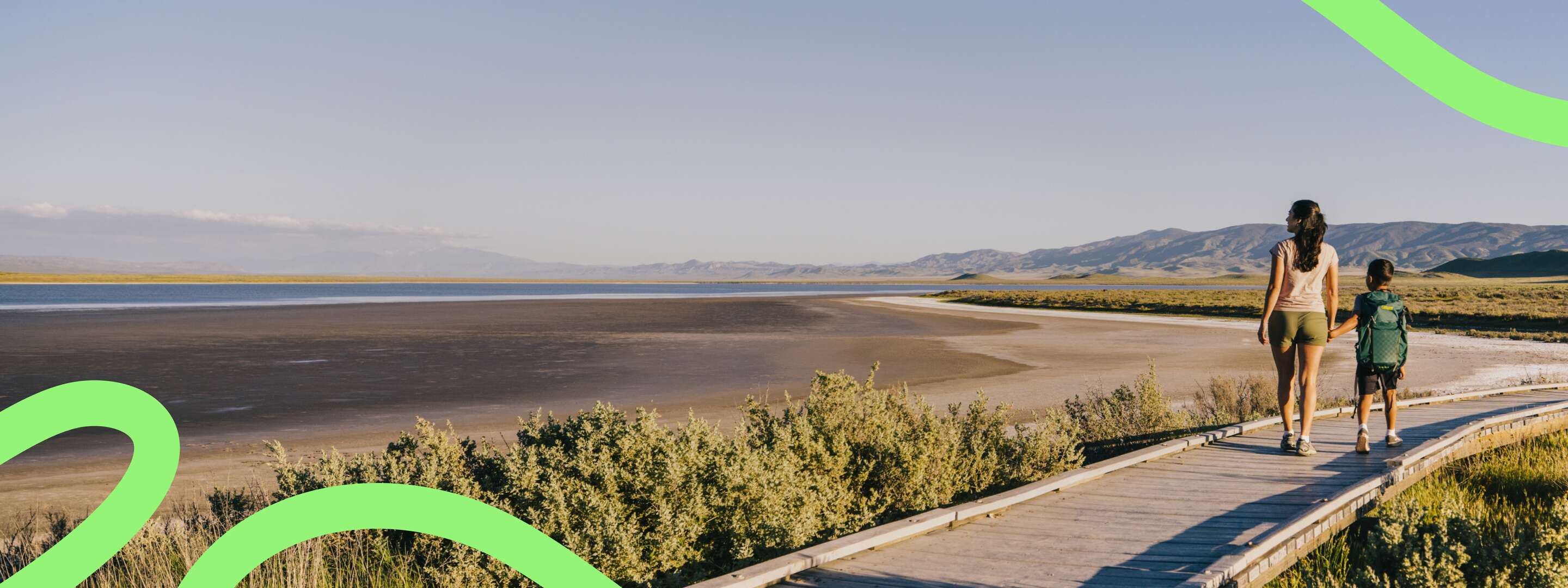 A mother and son walking across a wooden bridge that looks out onto sun-lit mountains and a far-reaching lake.