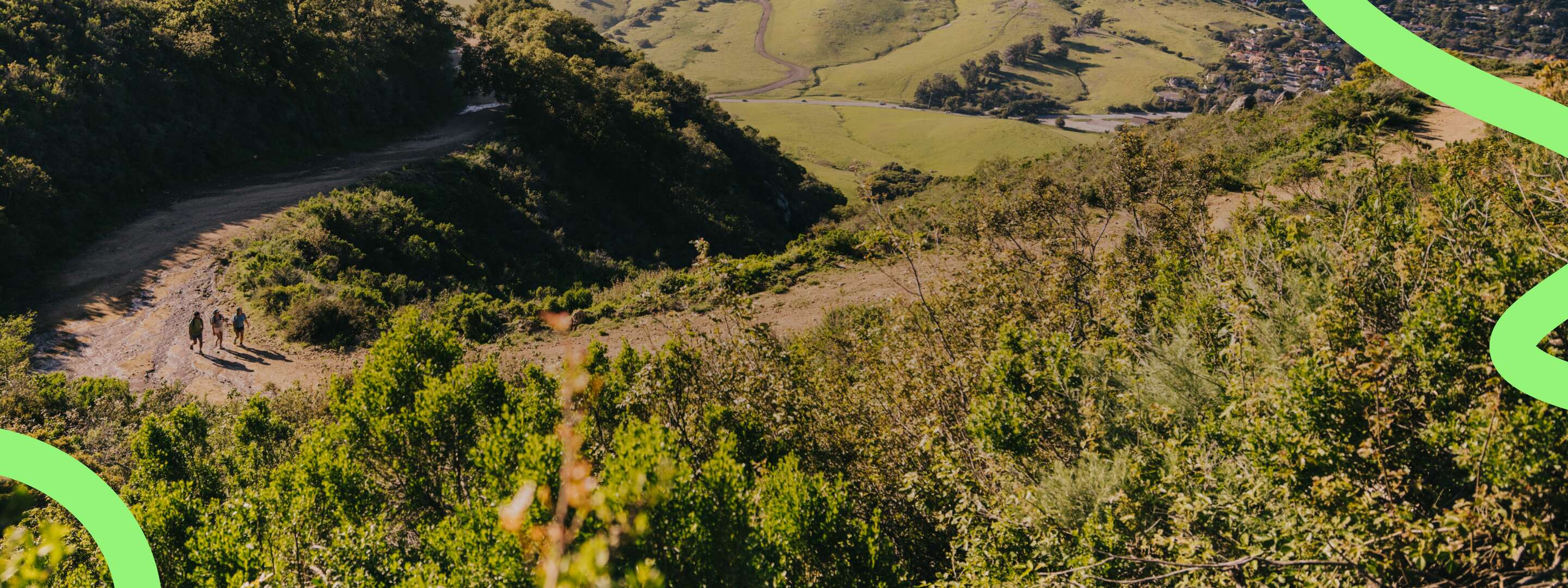 Three hikers walking on a trail, surrounded by sprawling green hills and sun-lit mountains.